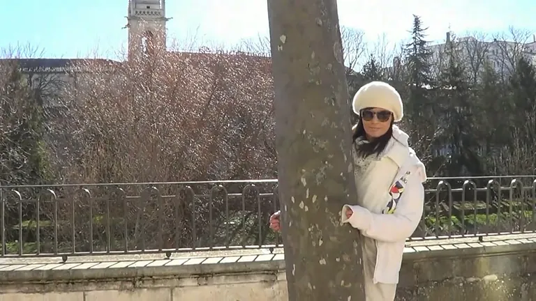 Mujer asomándose tras un árbol. Con bata blanca, sombrero y gafas de sol. Escenario al aire libre con edificios y árboles.