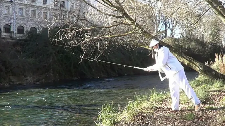 Una persona con traje blanco se adentra en un río con un palo largo. Edificio y árboles al fondo.