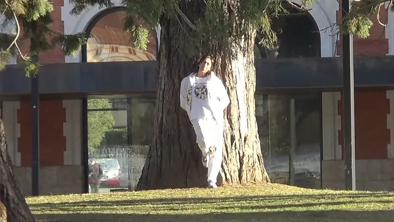 Una persona vestida de blanco se apoya contra un gran árbol en una zona cubierta de hierba, con un edificio visible al fondo.