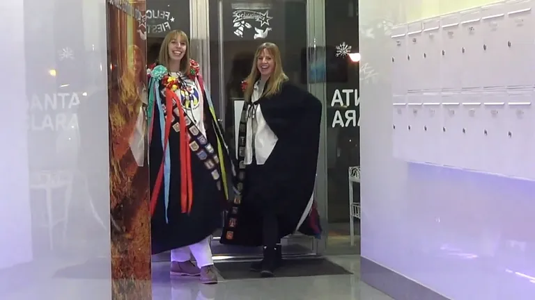 Dos mujeres con capas, una colorida con cintas, la otra negra, saliendo de un edificio con el nombre 