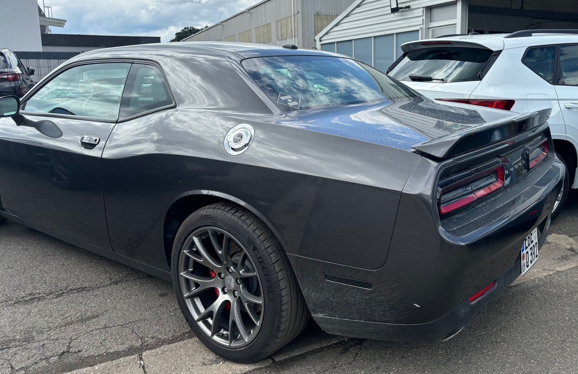 Dunkelgrauer Dodge Challenger auf einem Parkplatz, Seitenansicht, mit schwarzem Spoiler und roten Rücklichtern.
