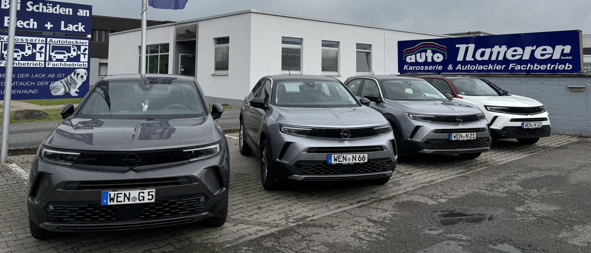 Vier neue graue SUVs parken vor einem Autohaus. Ein weißes Gebäude und ein Schild sind zu sehen.