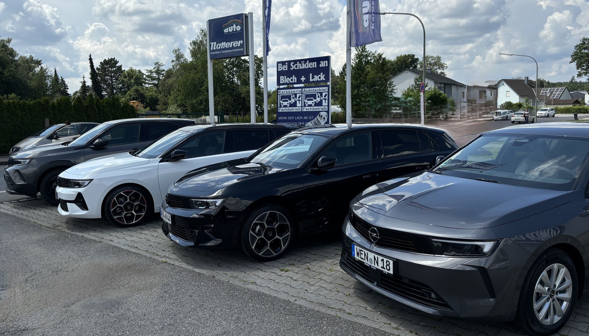 Vor einem Autohaus geparkte Autos in verschiedenen Farben. Schilder darüber und Gebäude im Hintergrund.