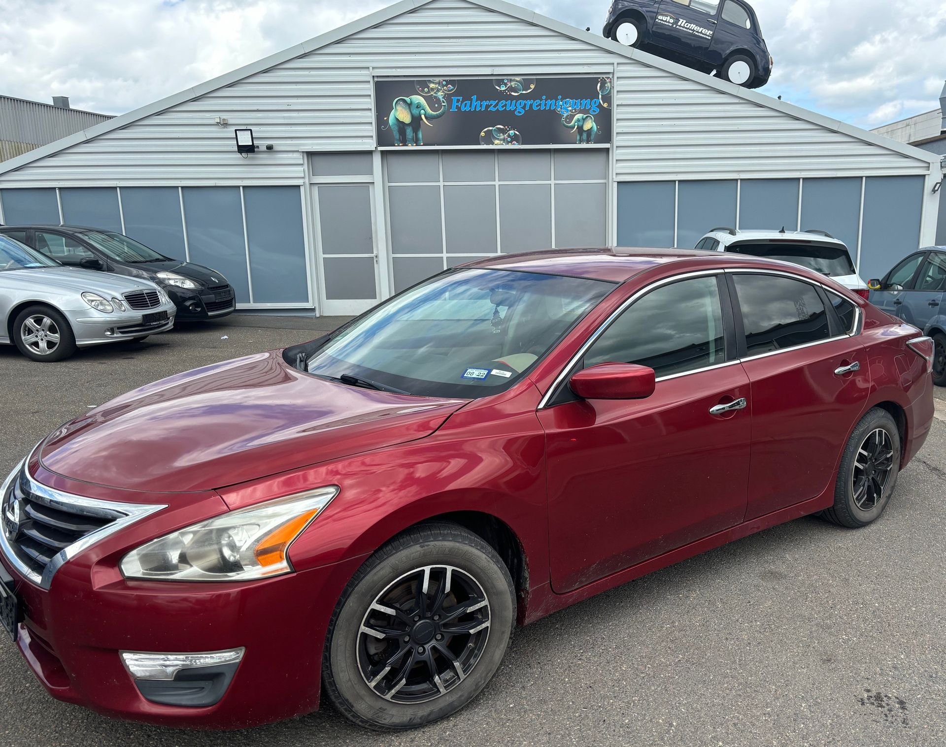 Roter Nissan Altima mit einem Kleinwagen auf dem Dach vor einem Autohaus geparkt.