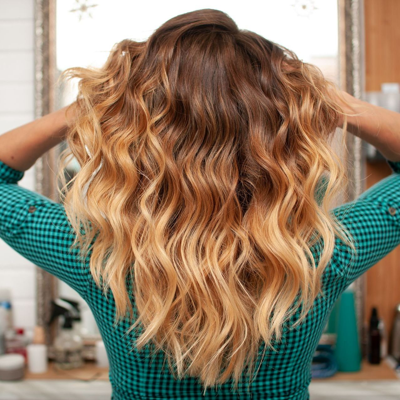 Femme de dos avec un haut vert et noir à carreaux qui se tient les cheveux avec un balayage blond chaud 