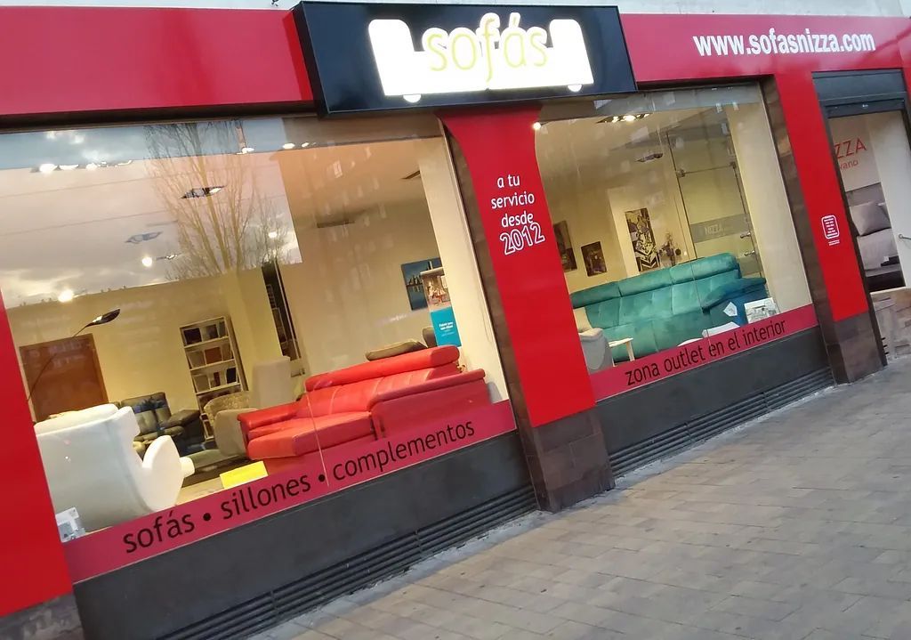 Fachada con letreros rojos y negros, grandes ventanales y sofás expuestos en el interior.
