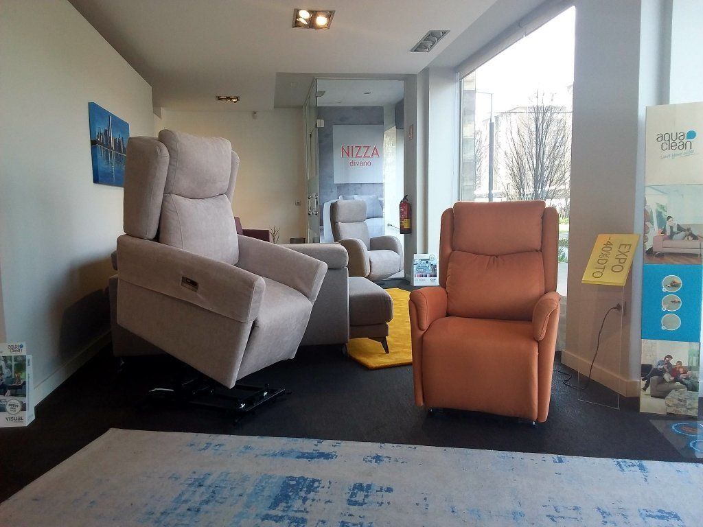 Sala de exposición de muebles con dos sillones reclinables, una silla gris y una silla naranja, sobre un suelo negro.