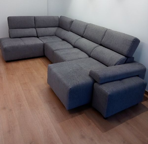 Sofá seccional gris con chaise longue y otomana sobre un suelo de madera clara en una habitación vacía.