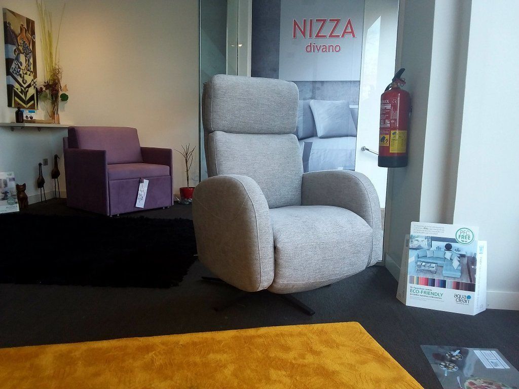 Sala de exposición con dos sillones reclinables grises, una silla morada y una alfombra negra sobre una alfombra amarilla.