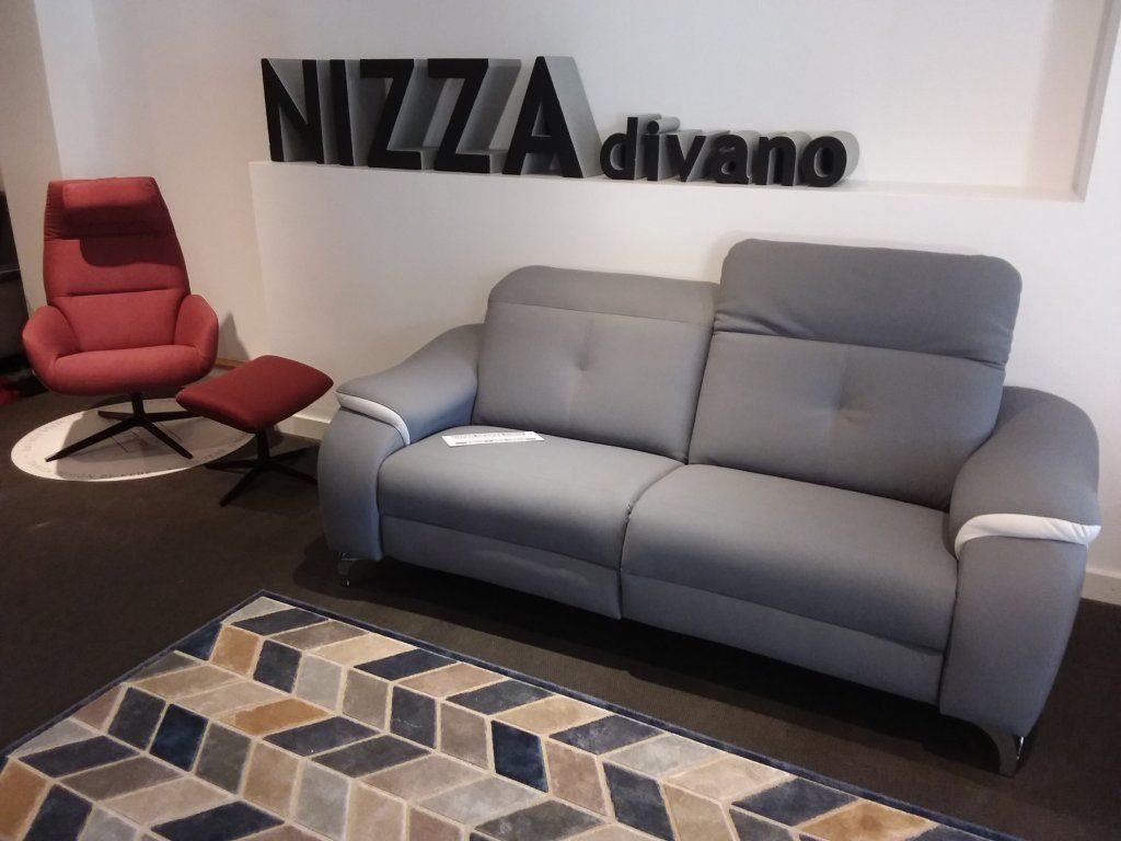 Sala de exposición con sofá gris, silla roja, alfombra estampada y letrero de Nizza divano en la pared.