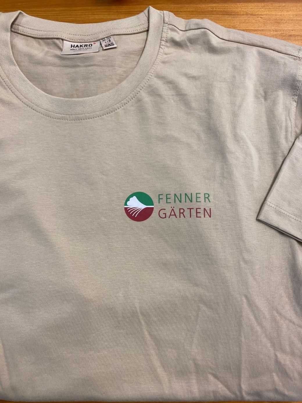 Auf einem Holztisch liegt ein weißes T-Shirt mit der Aufschrift „Fenner Garten“.