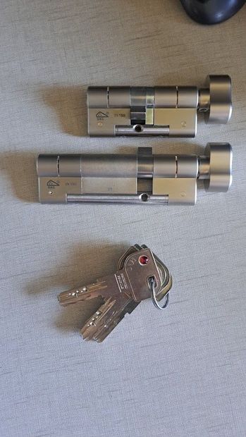 Dos cilindros de cerradura de puerta plateados con un pequeño llavero sobre una superficie beige.