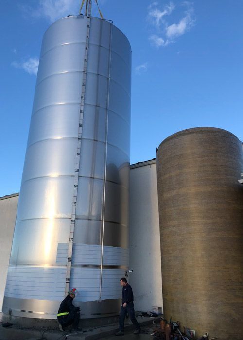 Dos hombres están de pie junto a un gran silo de acero inoxidable.