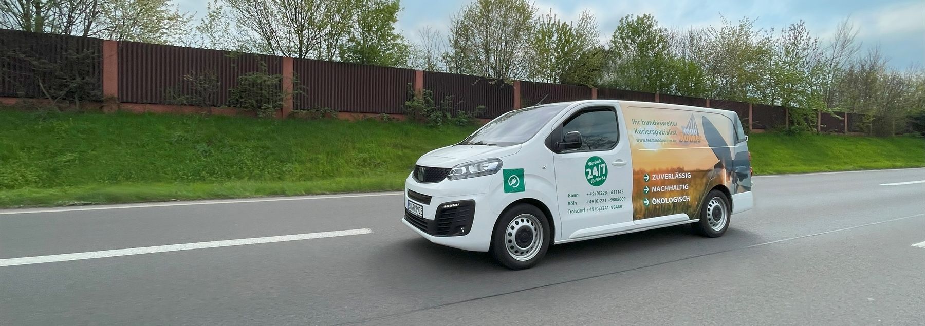Weißer Lieferwagen mit grüner und brauner Werbung auf einer Straße, vorbei an einem grünen Rasen und einem braunen Zaun.