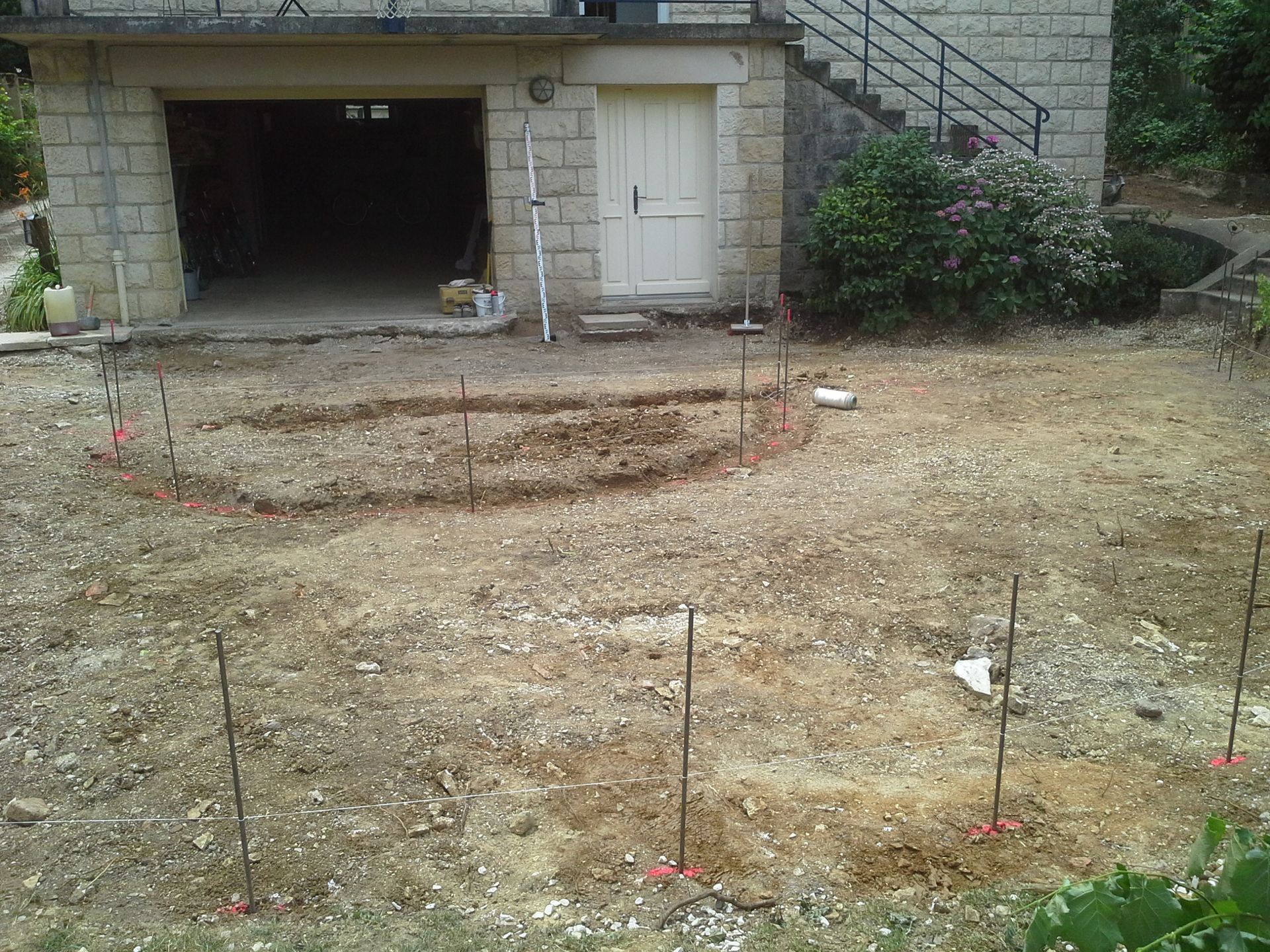 Terrain en terre délimité par des piquets, potentiellement pour aménagement paysager, devant un garage et un bâtiment en pierre.
