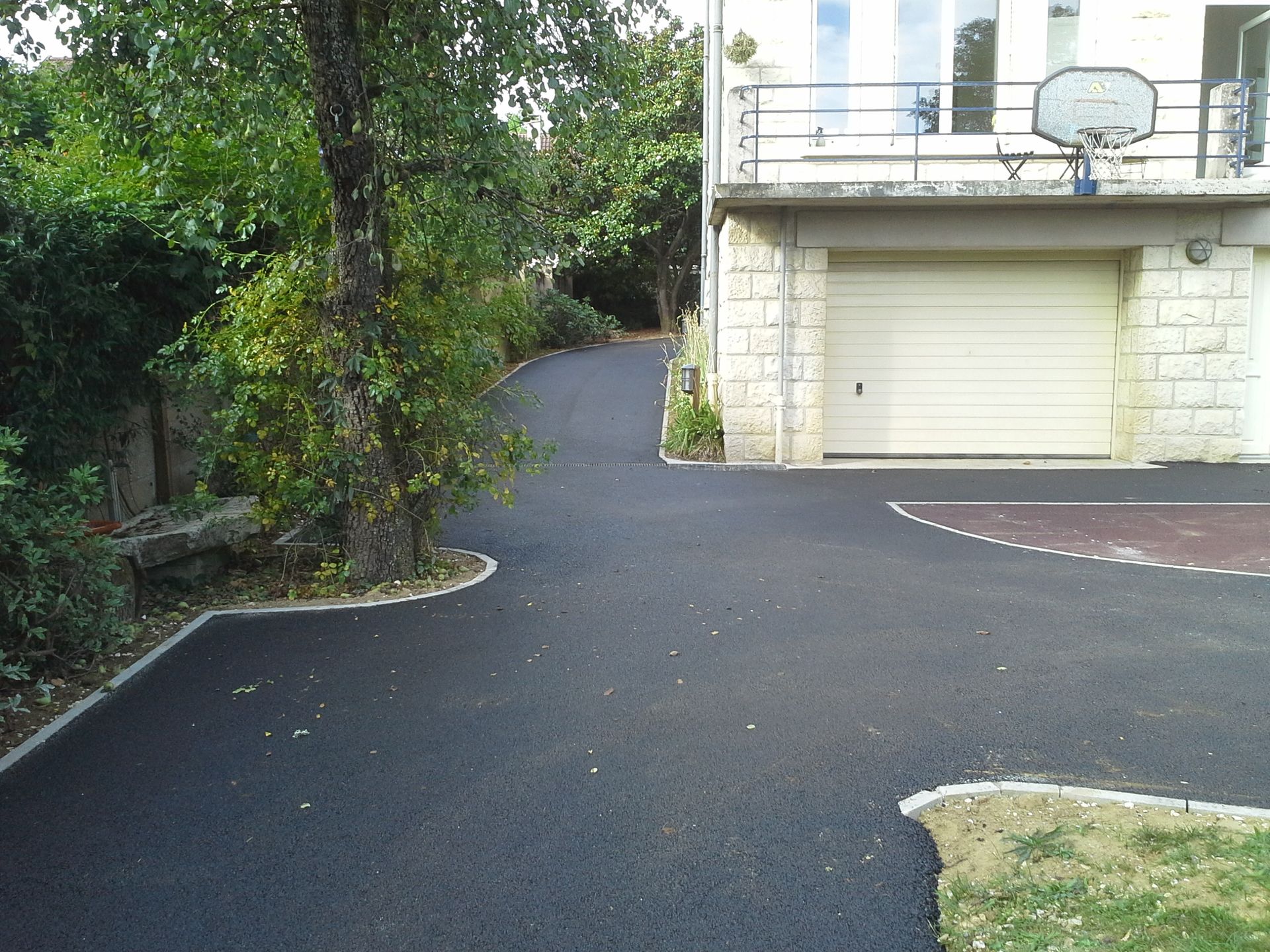 Une allée en asphalte noir serpente jusqu'à une maison avec un garage et un panier de basket, bordée d'herbe et d'arbres.