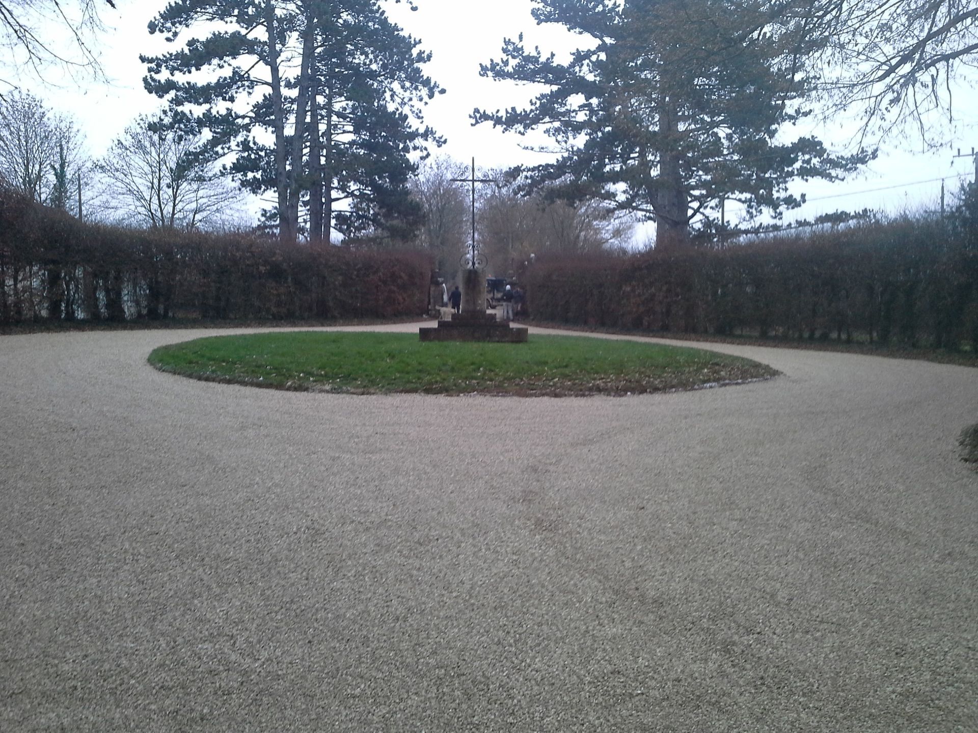 Chemin de gravier menant à une pelouse circulaire ornée d'un monument sombre. Arbres et buissons en arrière-plan.
