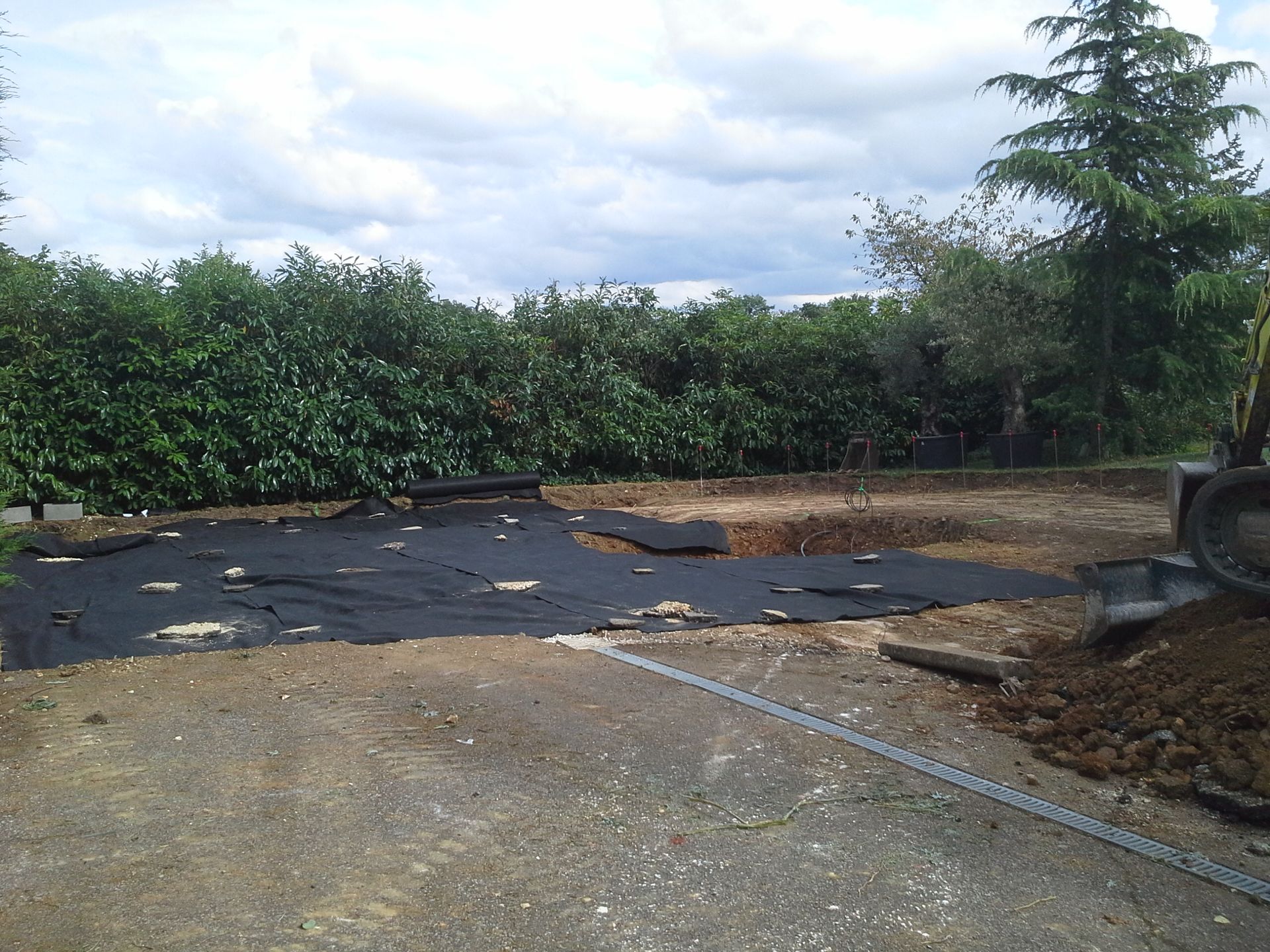 Une allée en gravier surmontée d'un écran anti mauvaises herbes noir. Des arbres et des broussailles bordent l'arrière-plan.