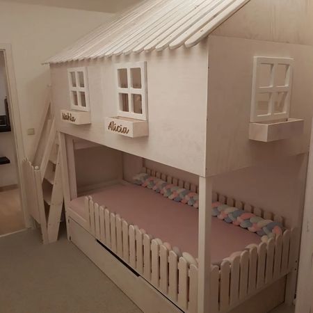 Ein weißes, hölzernes Etagenbett in Form eines Spielhauses mit schrägem Dach, Fensterläden und einem Lattenzaun als Sockel.