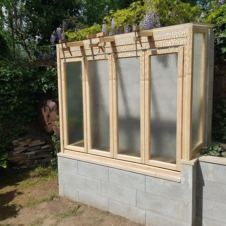 Inmitten eines Gartens thront eine Holzrahmenkonstruktion mit strukturierten Glaspaneelen auf einer Mauer aus Betonsteinen.