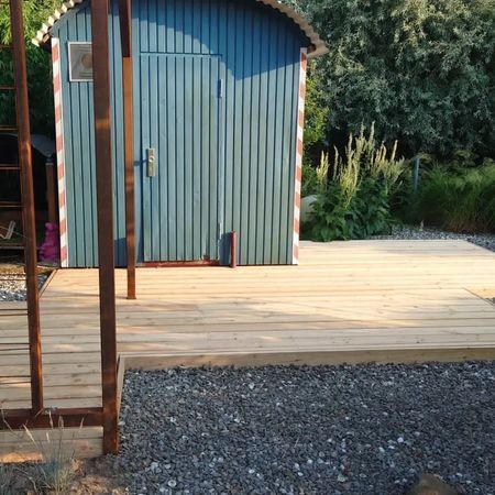 Vor einem blauen, bogenförmigen Geräteschuppen befindet sich eine neue Holzterrasse in einem Garten mit Kiesvordergrund.