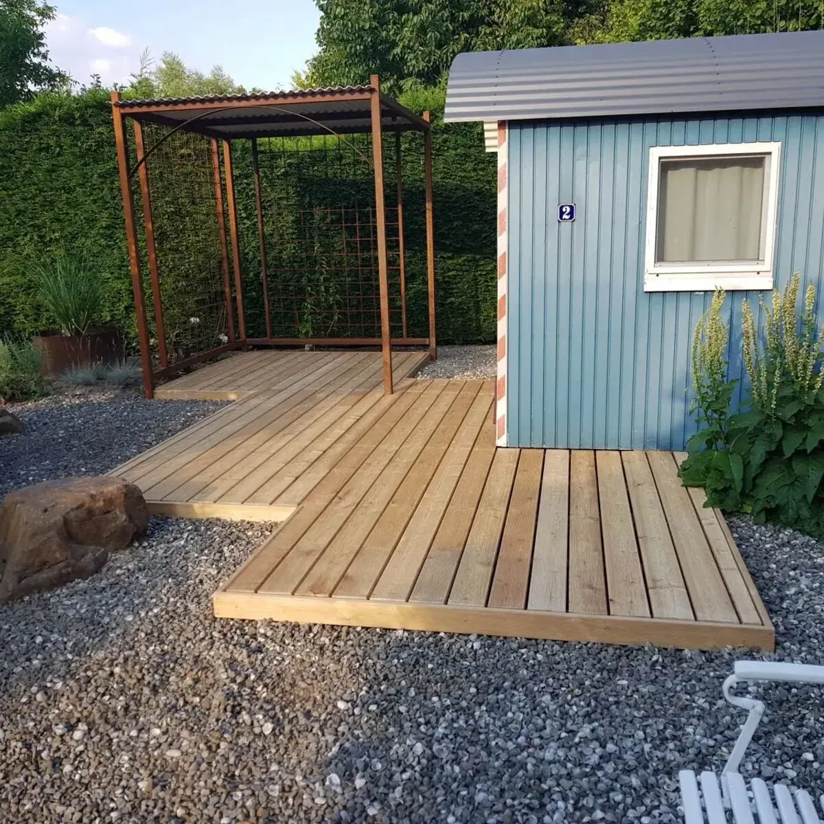 Ein blauer Wellblechschuppen mit Holzterrasse und passender Holzpergola auf Kiesgrund.