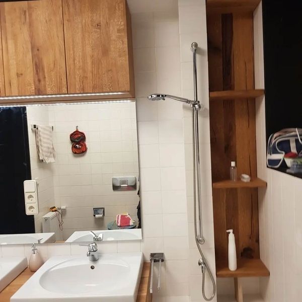 Ein Waschbecken, ein Spiegel und ein Duschkopf neben offenen Holzregalen an einer weiß gefliesten Wand.