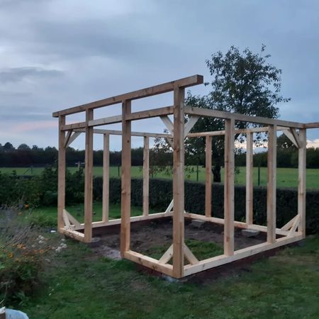 Eine im Bau befindliche Holzrahmenkonstruktion mit vertikalen Pfosten und diagonalen Stützen auf einer Rasenfläche.
