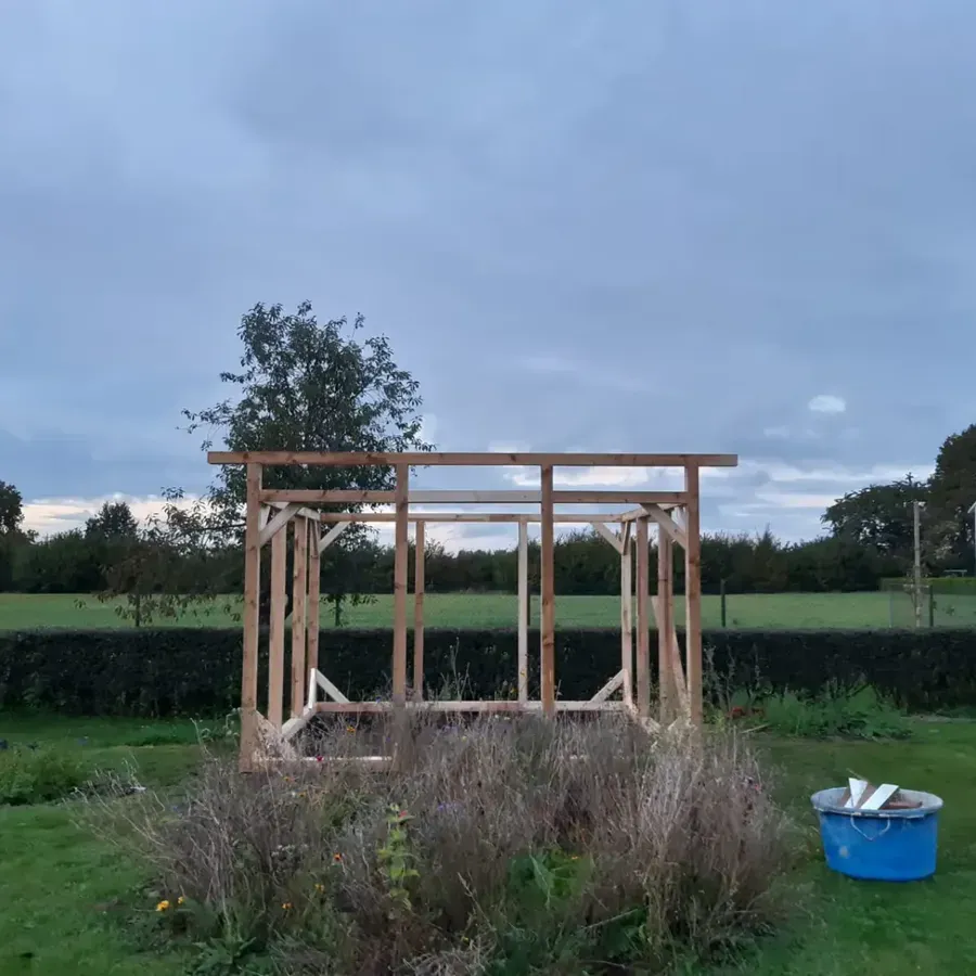 Auf einem grasbewachsenen Feld unter bewölktem Himmel steht das hölzerne Gerüst eines kleinen Schuppens.