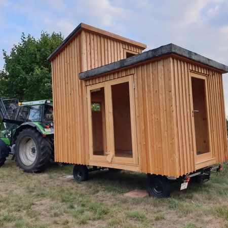 Auf einem grasbewachsenen Feld zieht ein grüner Traktor einen modernen, mobilen Holzschuppen
