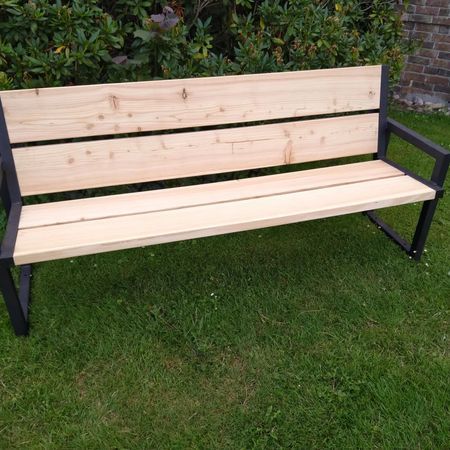 Eine Holzbank mit schwarzem Metallgestell, die auf einem grünen Rasen vor einem Gebüsch steht.