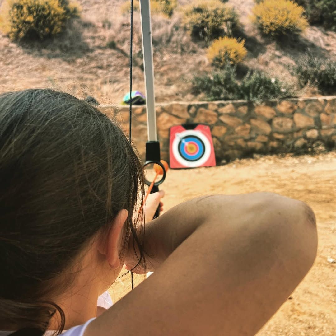 Persona apuntando con arco y flecha a una diana. La diana está frente a un muro de piedra, en un entorno soleado.