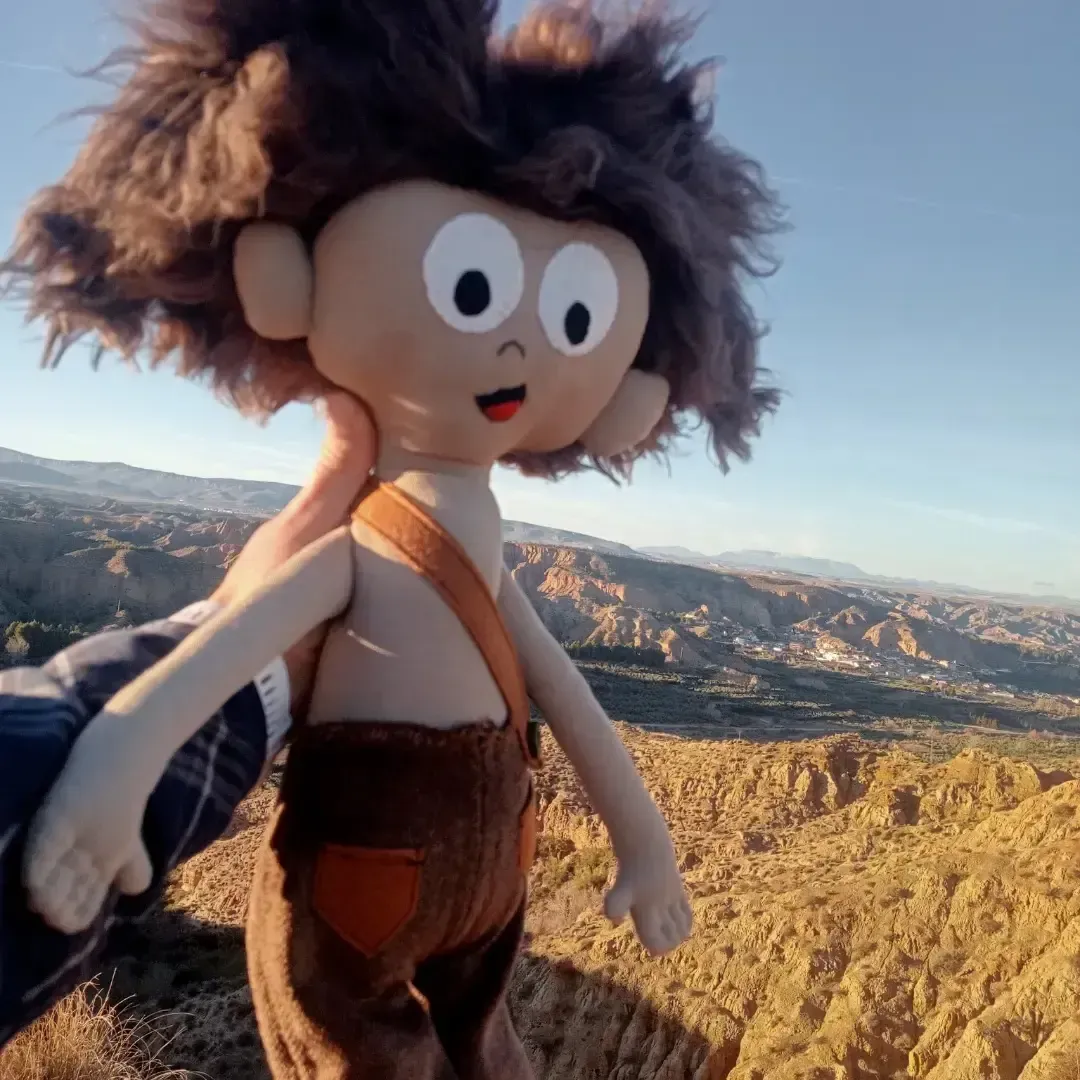 Figura de peluche sostenida en una montaña. Cabello y ojos castaños, y mono. Piel clara. Fondo montañoso.