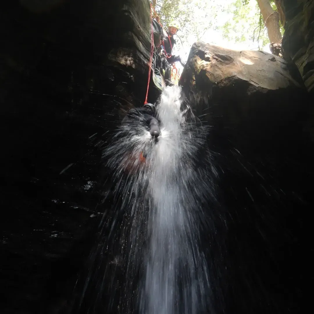 Persona descendiendo en rapel por una cascada en un estrecho cañón. El agua salpica a su alrededor. Luz brillante en la cima.