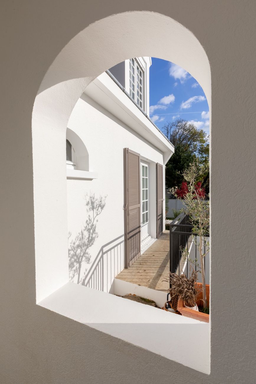 Un mur latéral en stuc clair d'une maison au toit pointu, partiellement masqué par un buisson et un arbre.