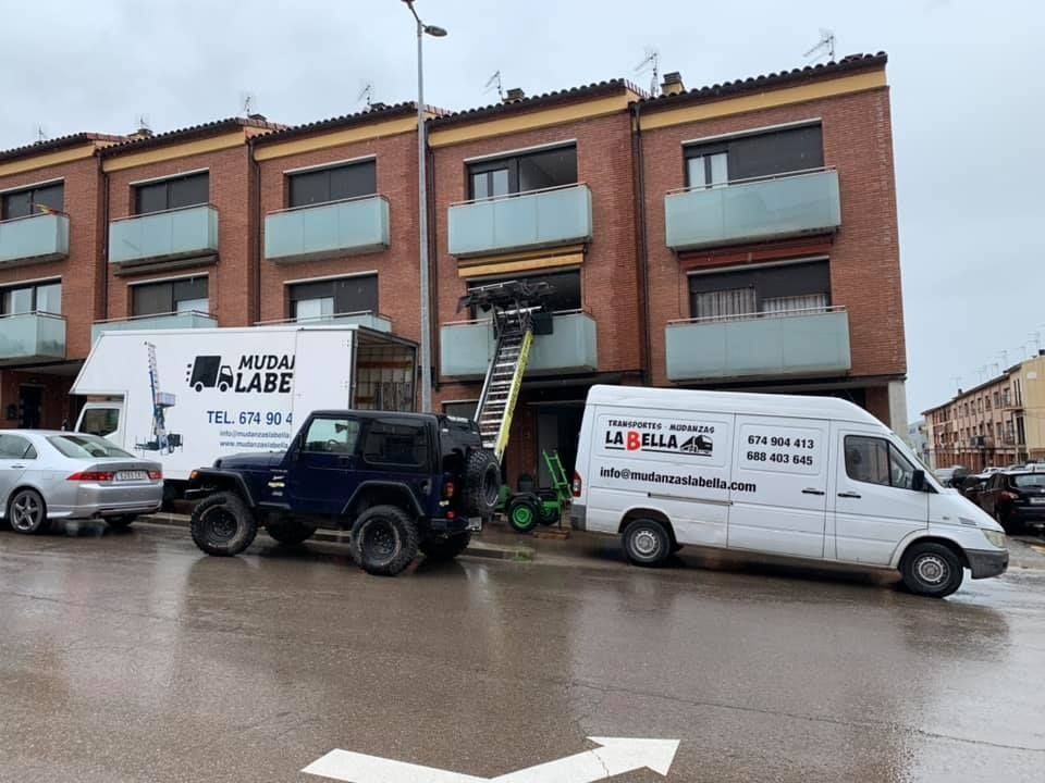 Camiones de mudanzas afuera de un edificio de ladrillos con balcones; trabajadores en una escalera.