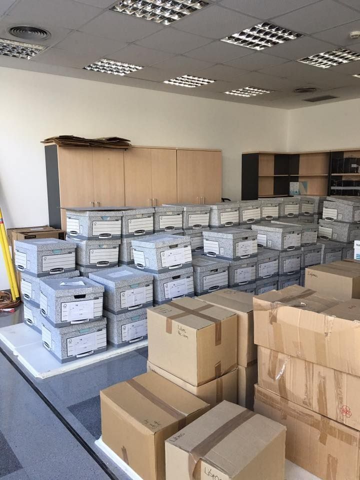 Filas de cajas de archivo grises y cajas de cartón apiladas en una habitación, bajo luz fluorescente. Los armarios se alinean en la pared del fondo.