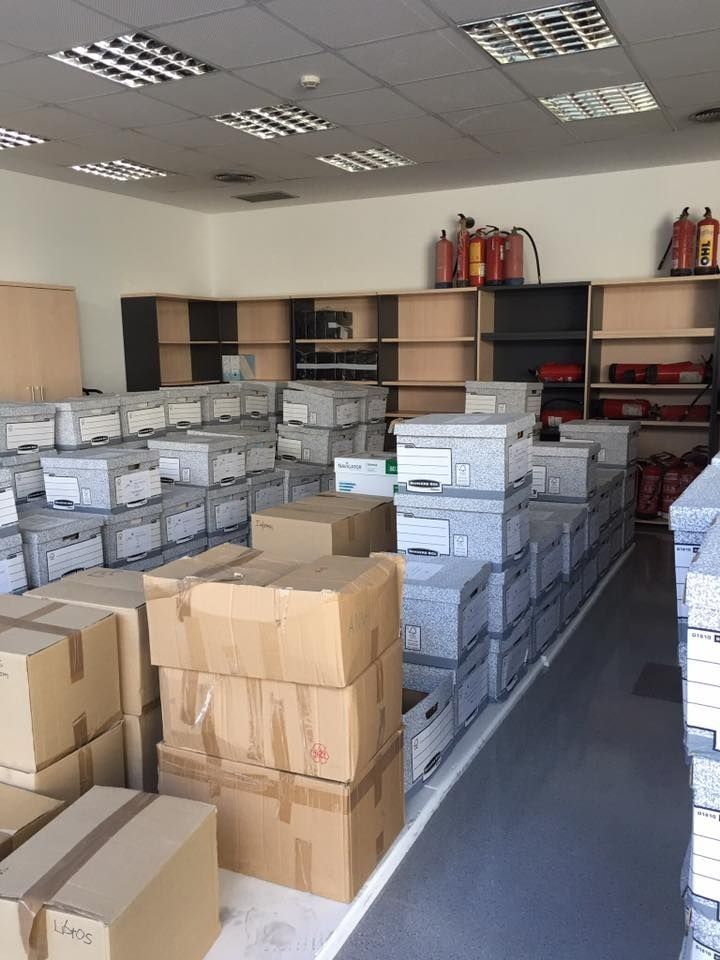 Trastero con pilas de cajas de cartón y archivadores grises. Estantes alinean la pared del fondo con extintores.