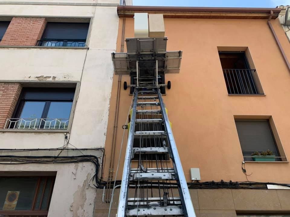 Ascensor en movimiento contra dos edificios, cajas en el andén. Escalera plateada, paredes color canela y crema.