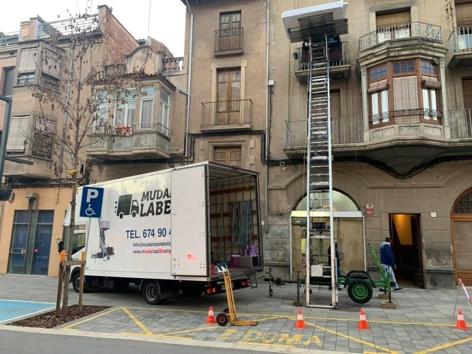 Camión de mudanzas con elevador junto al edificio; cargando muebles. Hay conos y una carretilla de mano.