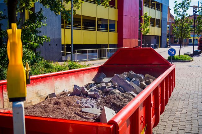 Am Straßenrand steht ein roter Müllcontainer voller Steine und Erde.