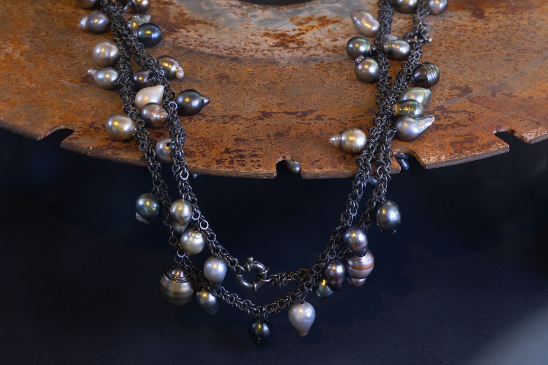 Dunkle Metallkette mit mehreren Strängen, Perlen und Verschluss, die auf einer rostigen, kreisförmigen Oberfläche ruht.