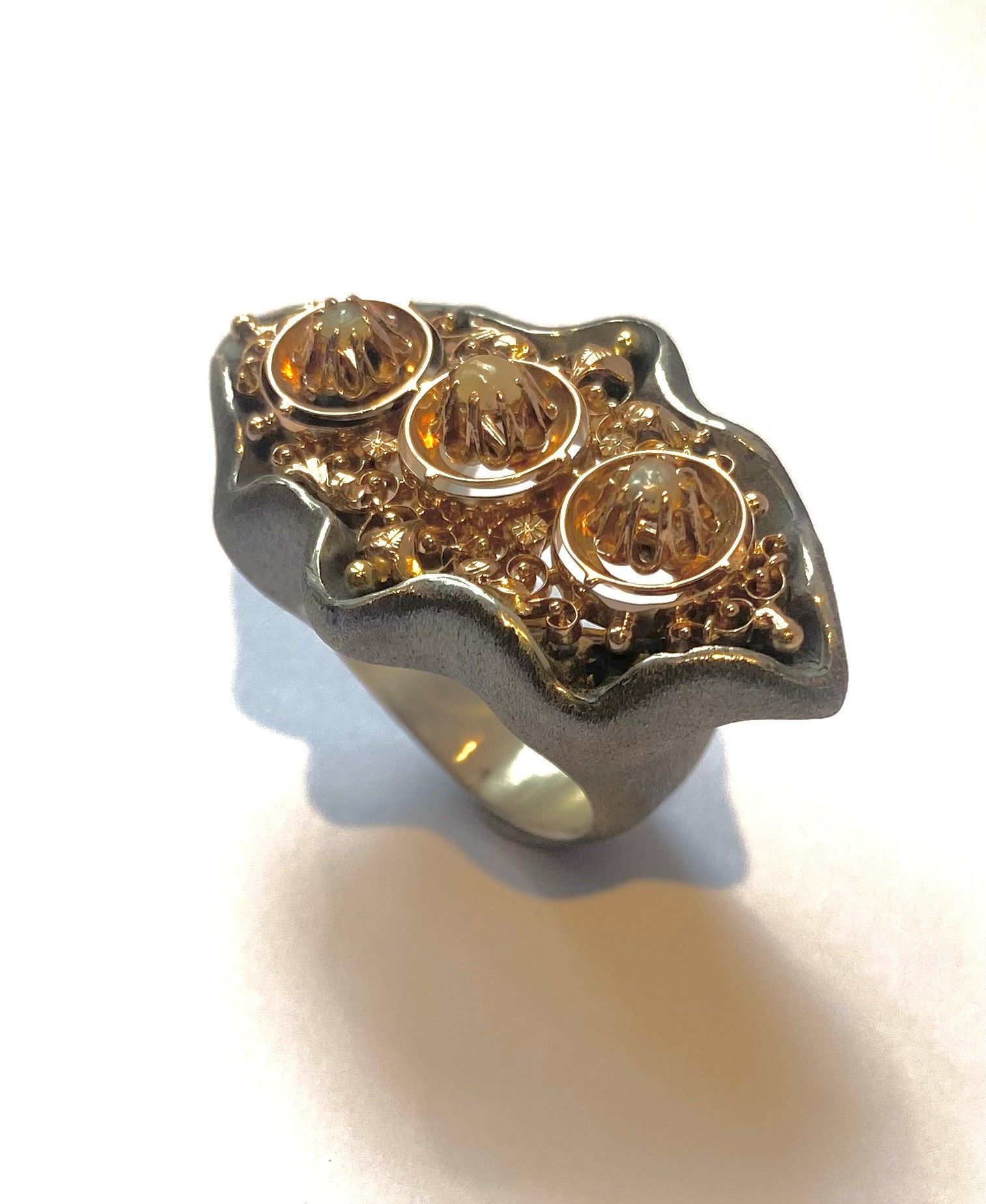 Silberner Ring mit dunklem, wellenförmigem Oberteil und drei goldfarbenen, kreisförmigen Details auf weißem Hintergrund