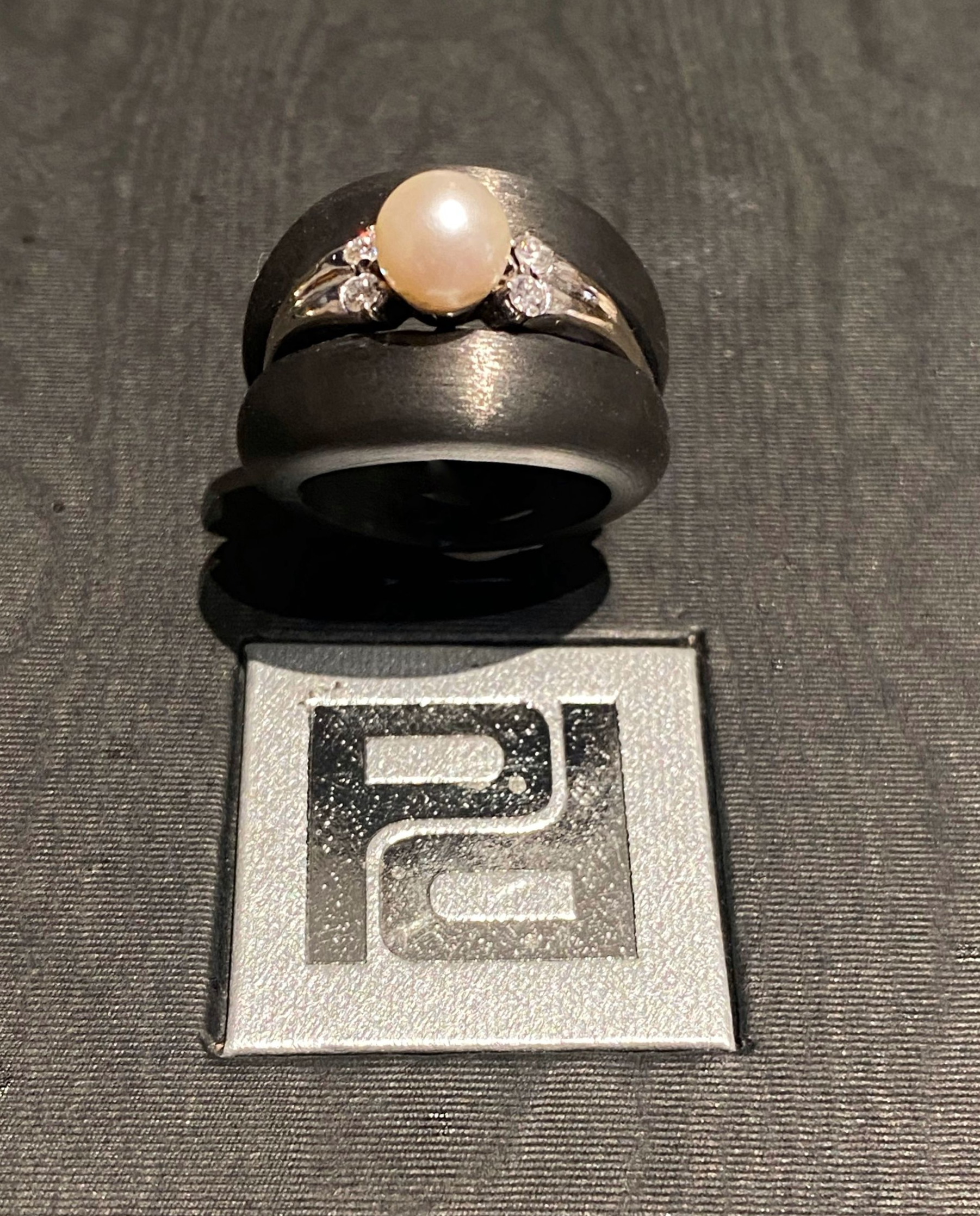 Ring mit Perle und Diamanten auf schwarzem Band. Darunter befindet sich das Logo mit der Aufschrift „PG“.