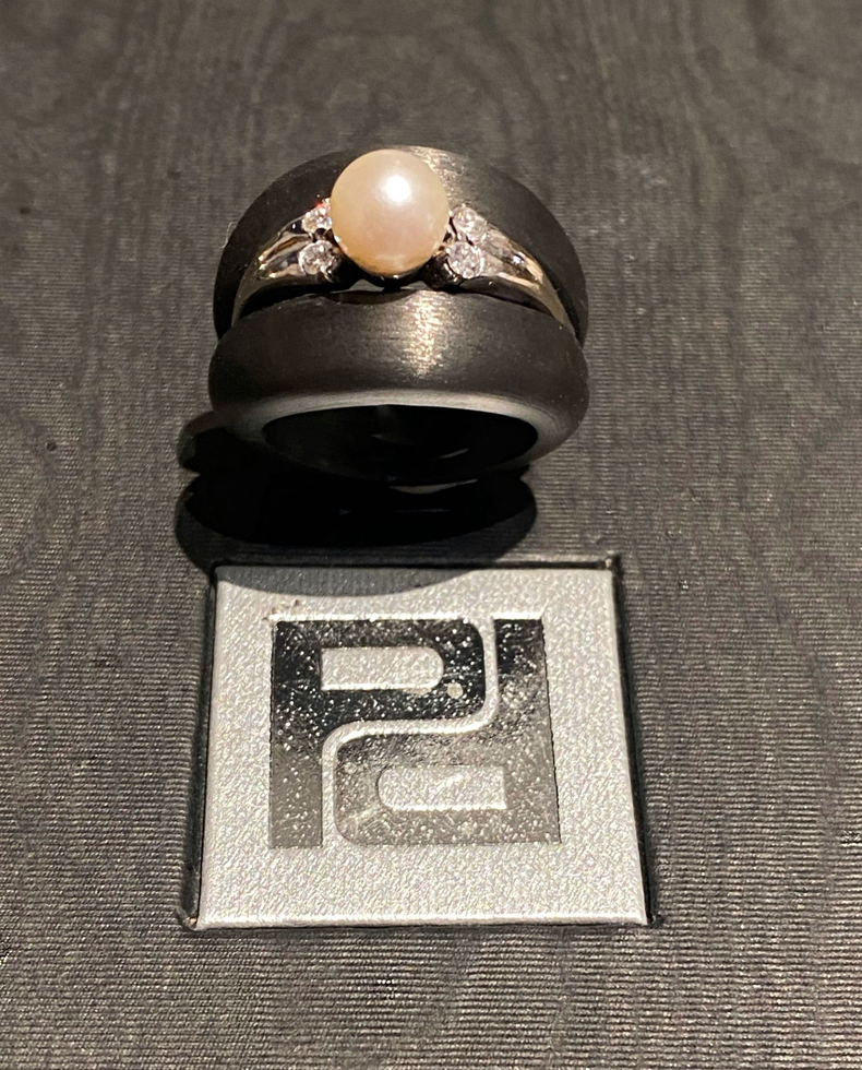 Ring mit Perle und Diamanten auf schwarzem Band. Darunter befindet sich das Logo mit der Aufschrift „PG“.