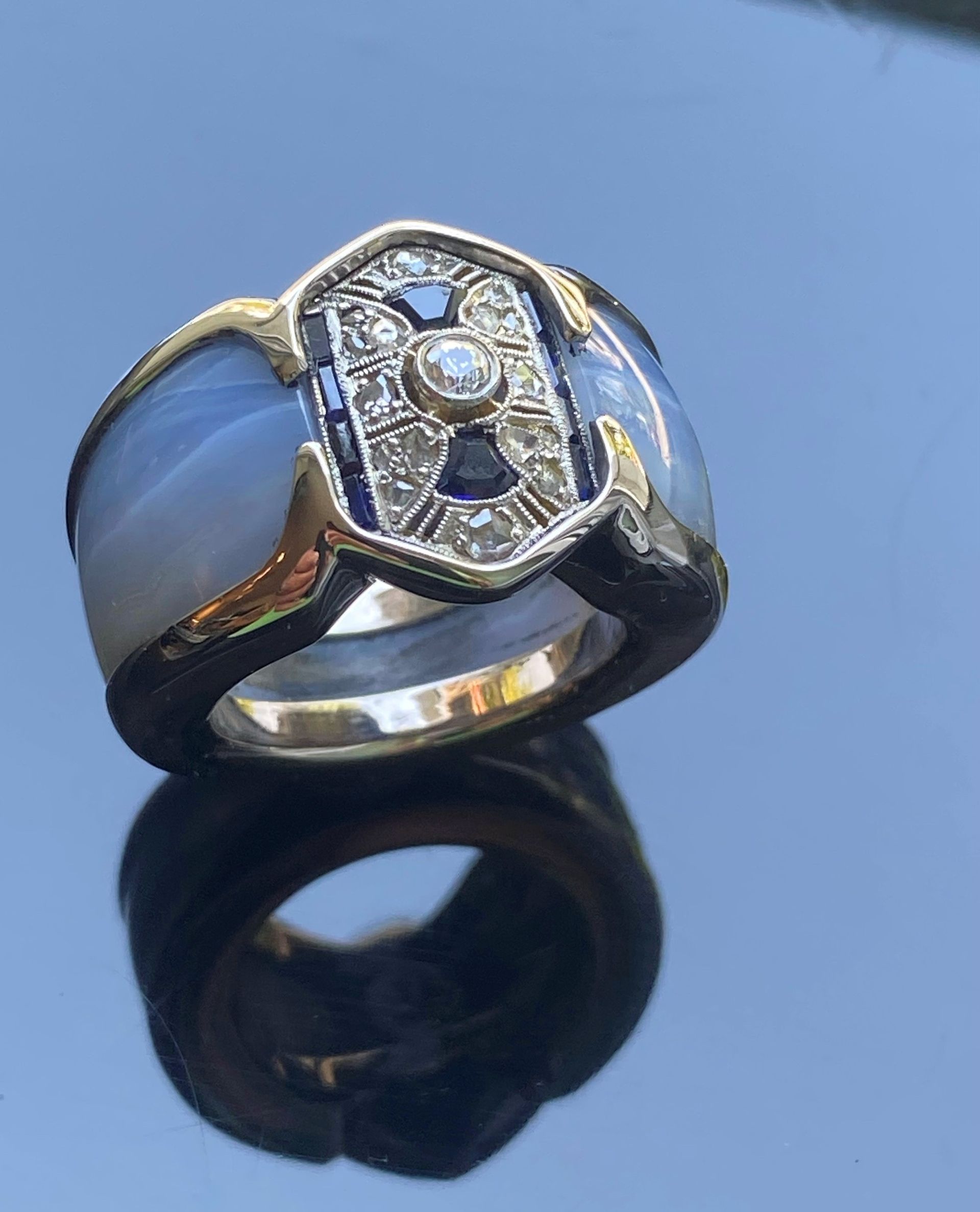 Schwarzer Ring mit silberner, rechteckiger Fassung und ovalem, blauem Stein, fotografiert auf einer reflektierenden Oberfläche