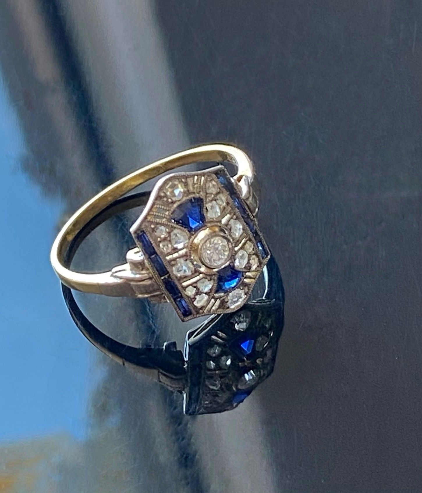 Goldring mit rechteckiger Fassung aus blauen und klaren Edelsteinen, die sich auf einer dunklen Oberfläche spiegeln