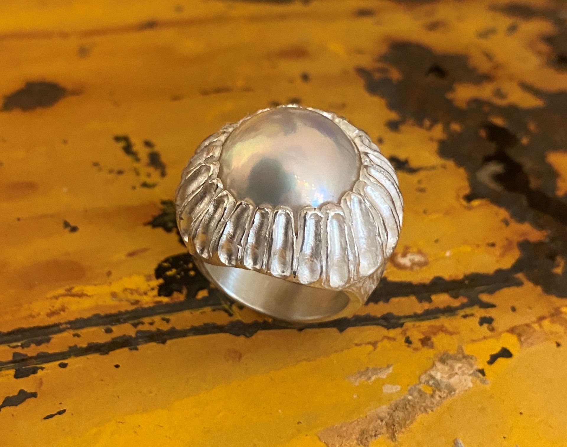 Silberner Ring mit einem runden, perlmuttartigen Mittelstein und einem geriffelten Ringband vor einem gelben Hintergrund.