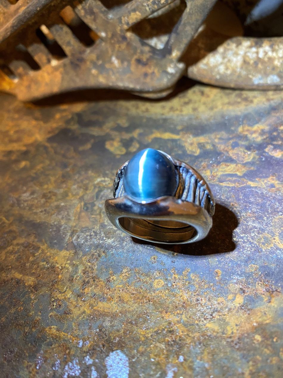 Silberring mit ovalem, blaugrauem Katzenaugenstein, eingefasst in eine rostige Metalloberfläche.
