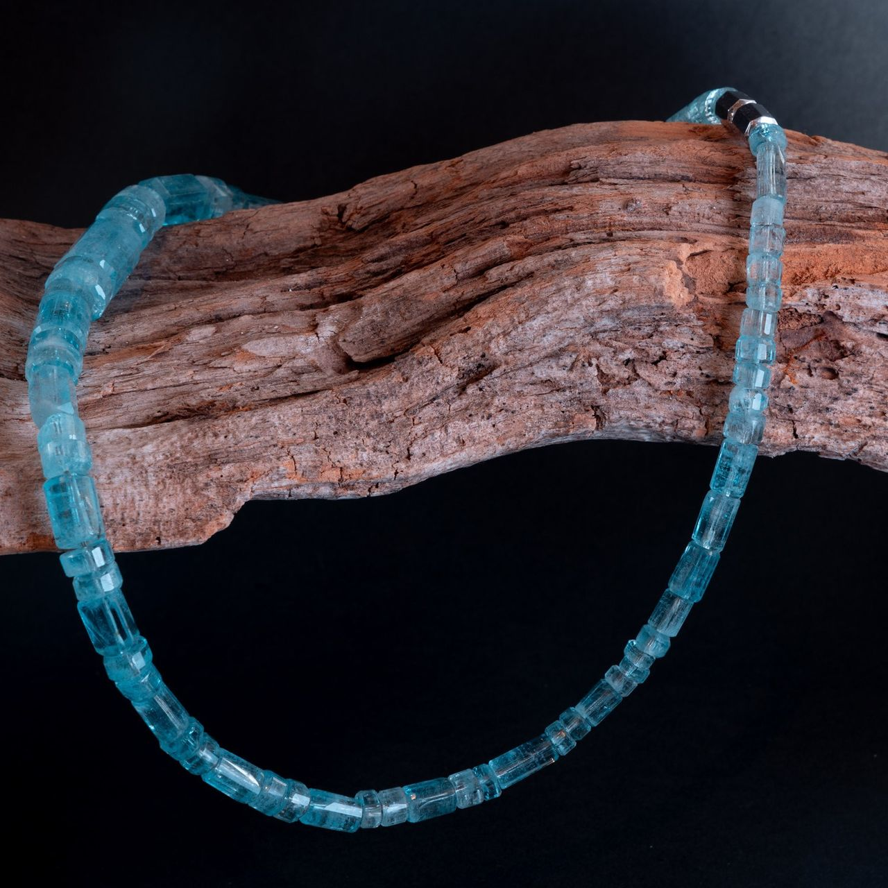 Eine blaue Perlenkette hängt über einem Stück verwittertem Holz vor schwarzem Hintergrund.
